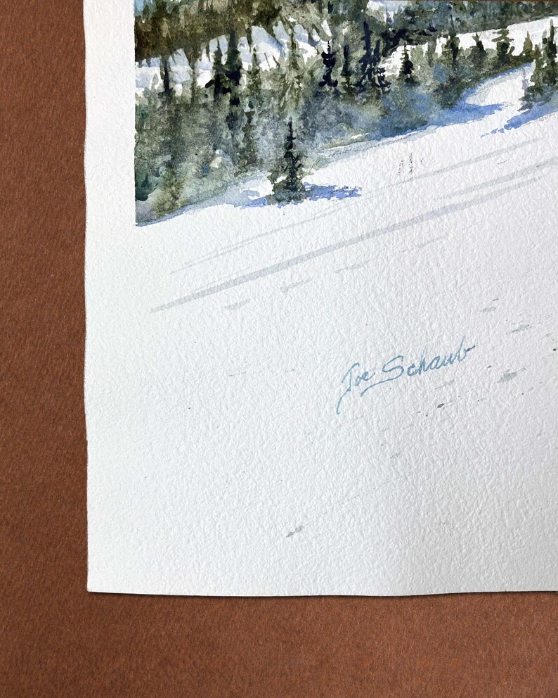 May include: Watercolor painting of a snow-covered mountain slope with evergreen trees. The artwork features a serene winter landscape with long shadows cast across the snow. The artist's signature, "Joe Schaub," is visible in the lower right corner.