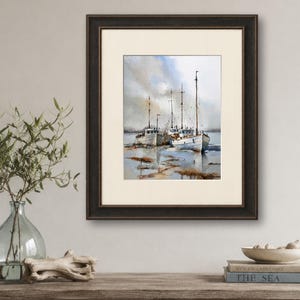 May include: A framed watercolor painting depicting three white fishing boats moored in shallow water under a cloudy sky. The artwork features muted tones of blue, gray, and brown, encased in a dark brown wooden frame with a cream-colored mat.