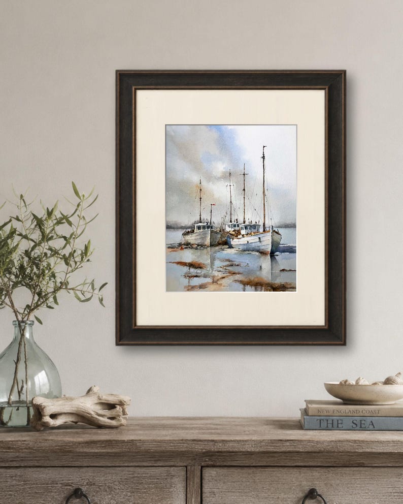 May include: A framed watercolor painting depicting three white fishing boats moored in shallow water under a cloudy sky. The artwork features muted tones of blue, gray, and brown, encased in a dark brown wooden frame with a cream-colored mat.