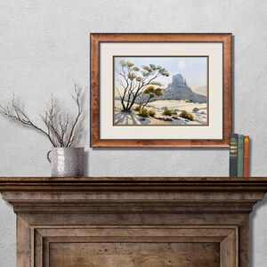 May include: A framed watercolor landscape painting depicts a desert scene with a mesa and sparse vegetation. The artwork is in a brown wooden frame and hangs above a wooden mantel. A decorative vase with branches and books are also on the mantel.