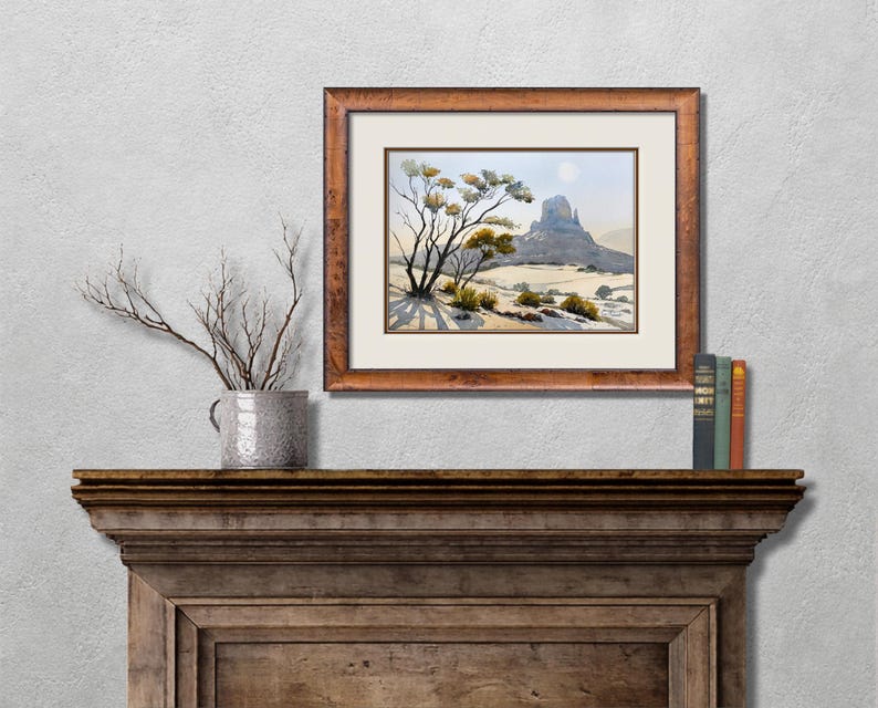 May include: A framed watercolor landscape painting depicts a desert scene with a mesa and sparse vegetation. The artwork is in a brown wooden frame and hangs above a wooden mantel. A decorative vase with branches and books are also on the mantel.