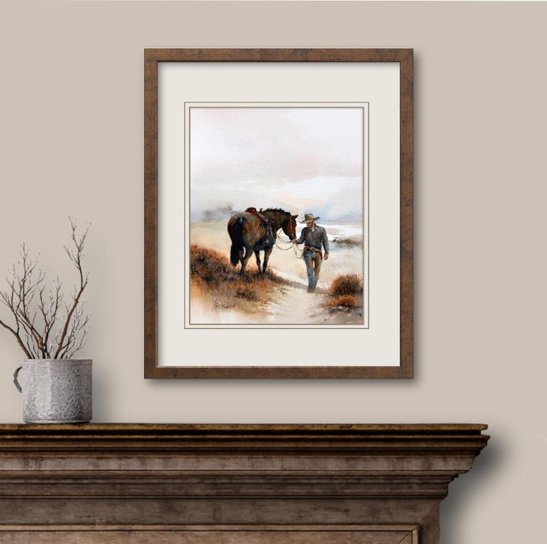 May include: Watercolor painting of a cowboy leading a horse on a path. The artwork is framed in a dark brown wooden frame with a white mat. The scene depicts a serene outdoor setting with muted colors.