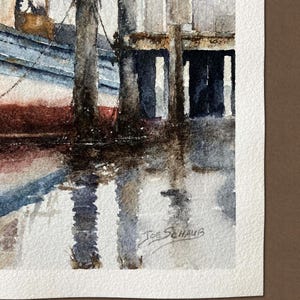 May include: A watercolor painting depicting a blue and white boat docked beside weathered wooden pilings. The scene features dark reflections in the water and the signature Joe Schaub in the bottom right corner on textured white paper.