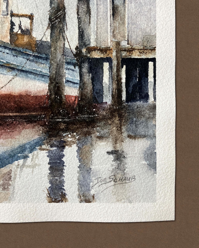 May include: A watercolor painting depicting a blue and white boat docked beside weathered wooden pilings. The scene features dark reflections in the water and the signature Joe Schaub in the bottom right corner on textured white paper.