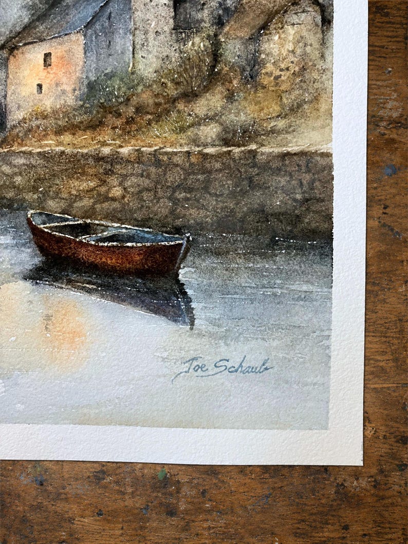 Puede incluir: Una pintura de acuarela que representa un peque&ntilde;o bote marr&oacute;n flotando en aguas tranquilas cerca de un muro de piedra y un edificio con luz c&aacute;lida en las ventanas. La obra presenta un borde de papel texturizado y la firma Joe Schaub en la esquina inferior derecha.