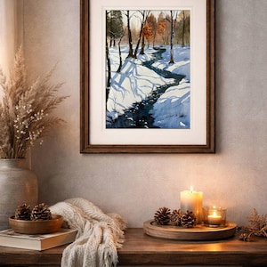 May include: A framed watercolor painting depicts a snowy winter landscape with a stream winding through a forest. The painting is displayed on a wooden table with candles, pine cones, and a decorative vase.