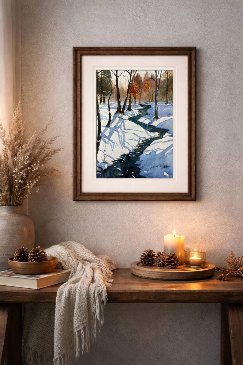 May include: A framed watercolor painting depicts a snowy winter landscape with a stream winding through a forest. The painting is displayed on a wooden table with candles, pine cones, and a decorative vase.
