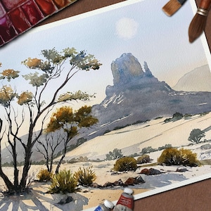 May include: Watercolor painting of a desert landscape with a large rock formation and sparse vegetation. The artwork features a palette of muted blues, browns, and greens. Paint tubes and brushes are visible, suggesting an artist's studio.