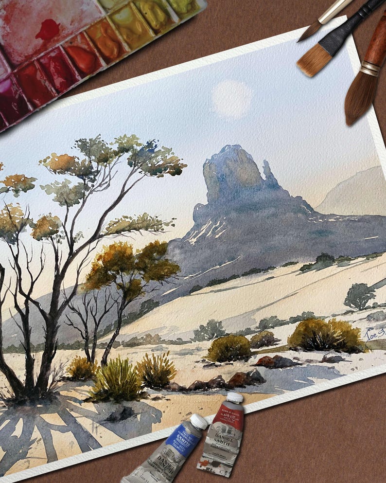 May include: Watercolor painting of a desert landscape with a large rock formation and sparse vegetation. The artwork features a palette of muted blues, browns, and greens. Paint tubes and brushes are visible, suggesting an artist's studio.