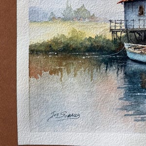May include: Watercolor painting of a lakeside scene. A light blue building with a red roof sits on stilts. A small blue boat is tied to the building. The water reflects the sky and the surrounding foliage. Signed "Joe Schaub".