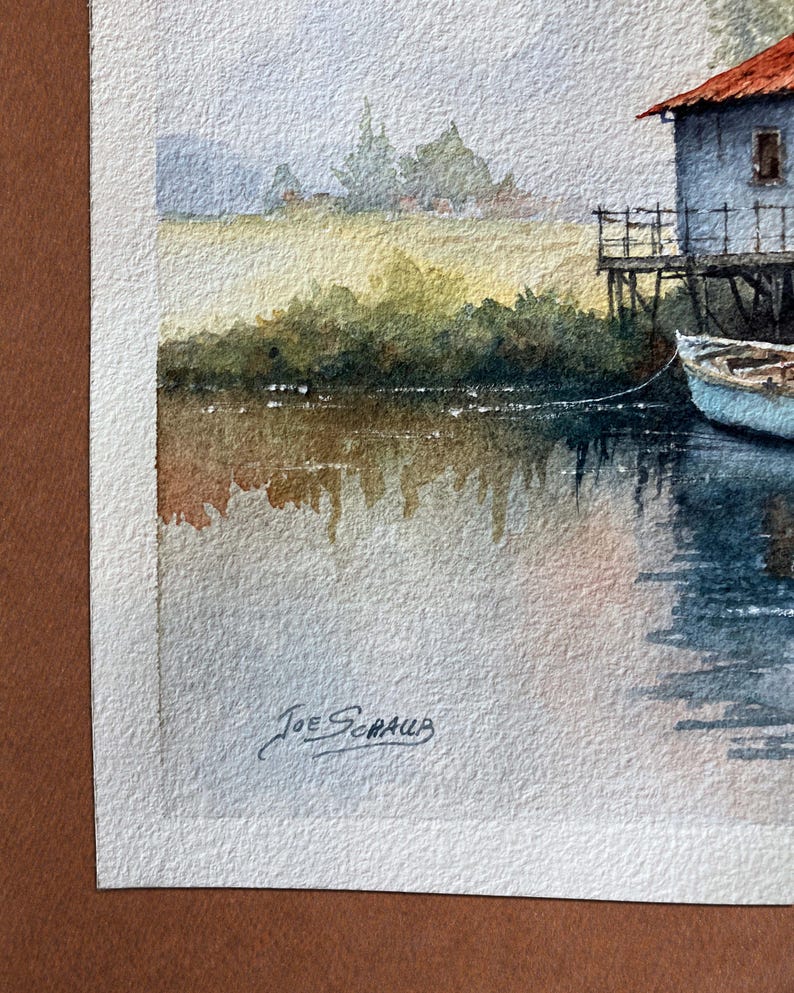 May include: Watercolor painting of a lakeside scene. A light blue building with a red roof sits on stilts. A small blue boat is tied to the building. The water reflects the sky and the surrounding foliage. Signed "Joe Schaub".
