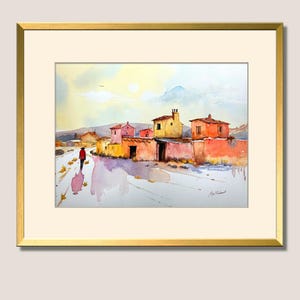 May include: Watercolour painting depicting a village scene with vibrant buildings and a figure in a red coat on a path. The artwork is framed in a gold frame with a cream-coloured mount.