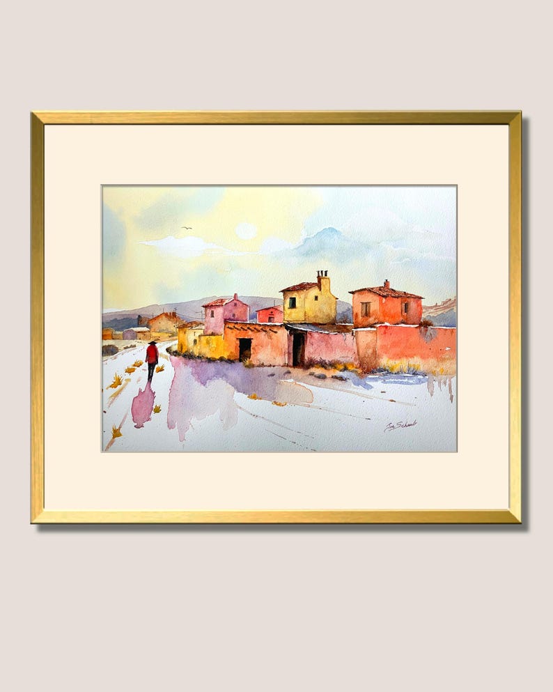 May include: Watercolour painting depicting a village scene with vibrant buildings and a figure in a red coat on a path. The artwork is framed in a gold frame with a cream-coloured mount.
