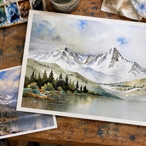May include: A watercolor painting on paper depicting a snow-capped mountain range rising above a dark forest and a calm lake. The scene features soft blue and gray sky tones, white snow on jagged peaks, and deep green pine trees near the water.