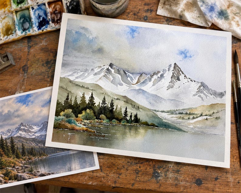 May include: A watercolor painting on paper depicting a snow-capped mountain range rising above a dark forest and a calm lake. The scene features soft blue and gray sky tones, white snow on jagged peaks, and deep green pine trees near the water.