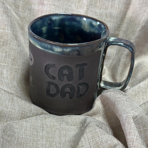 May include: A handmade ceramic mug with a dark brown exterior and a blue interior. The mug features the embossed text "CAT DAD" on its side. The handle and rim have a blue glaze, and the mug sits on a neutral-colored fabric.