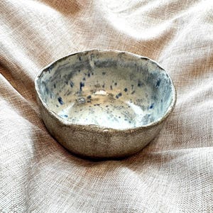 Op de afbeelding: Een handgemaakte keramische kom met een ruwe, getextureerde buitenkant. De binnenkant heeft een gespikkelde blauw-witte glazuur. De kom heeft een onregelmatige, organische vorm, wat suggereert dat het een uniek stuk aardewerk is.