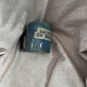 Peut inclure: Une tasse en céramique bleue avec une anse noire et le texte "BROKE ON PAYDAY" en noir. La tasse a une texture tachetée et est posée sur un fond en tissu de couleur neutre.