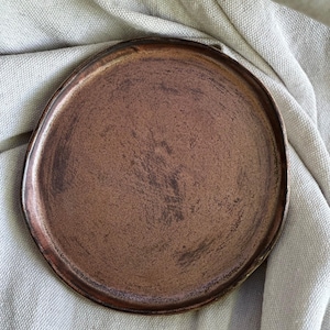 May include: A handcrafted, circular, copper-toned ceramic plate with a gently uneven rim. The plate features a textured finish and is displayed on a neutral linen cloth. The plate's colour ranges from light to dark copper.
