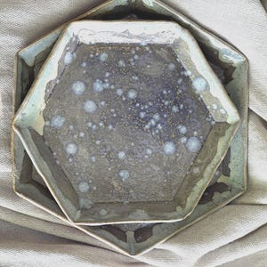 May include: Two hexagonal ceramic plates, one inside the other. The plates have a speckled, mottled surface in shades of grey, blue, and white. The edges are a lighter green-grey. The plates are on a neutral-coloured fabric.