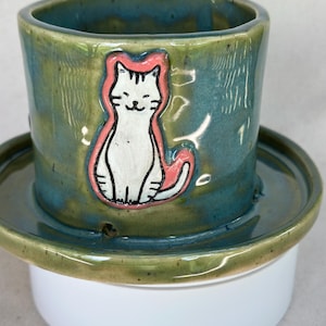 May include: A cylindrical ceramic cup with a glossy blue-green glaze featuring a raised, hand-painted white cat illustration outlined in pink. The cup sits on a matching wide-rimmed saucer, showcasing a smooth, reflective finish and detailed cat features.