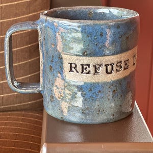 Puede incluir: Taza de cerámica hecha a mano con un esmalte azul y moteado. La taza presenta un asa rectangular y una banda con la palabra "REFUSE" en letras en relieve. La taza está sobre una superficie marrón.
