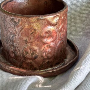 May include: A handcrafted, cylindrical ceramic planter with a matching saucer. The planter has a textured surface with a repeating floral pattern and a rich, reddish-brown color. The interior and saucer have a metallic sheen.