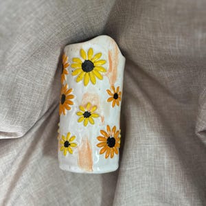 May include: A white ceramic vase with a cylindrical shape, adorned with hand-painted sunflowers in yellow and orange, each with a black center. The vase has a slightly textured surface and is set against a neutral fabric backdrop.