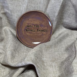 Peut inclure: Une petite coupelle ronde en céramique avec une finition brillante. La coupelle est de couleur marron foncé avec des reflets marron clair et violets. Les mots "ALL THE SMALL THINGS" sont en relief sur la surface. La coupelle est posée sur un tissu texturé de couleur neutre.