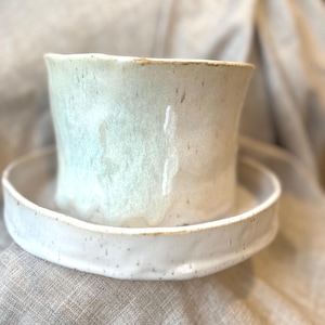 May include: A handmade ceramic planter with a matching saucer. The planter is a light cream color with a slightly flared shape and a textured surface. The saucer is round and has a slightly raised edge. The set is ideal for small plants.