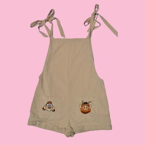 May include: A beige overall romper with adjustable straps and two embroidered cartoon animal faces on the front pockets. The animal faces are a brown and orange warthog and a brown and orange meerkat.