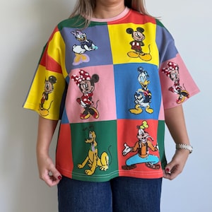 May include: A colorful patchwork t-shirt featuring various Disney cartoon characters. The shirt has a square pattern with characters like Mickey Mouse, Minnie Mouse, Donald Duck, and Goofy. The shirt is a mix of colors including yellow, pink, blue, green, and red.