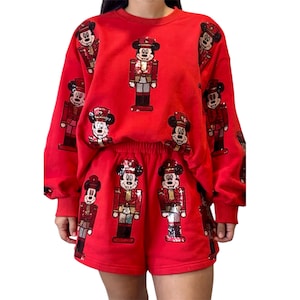 May include: A bright red long-sleeve sweatshirt and matching shorts set featuring a repeating sequined pattern of Mickey Mouse dressed as a nutcracker. The sequins are multicolored, adding texture and shine to the red fabric of the garments.