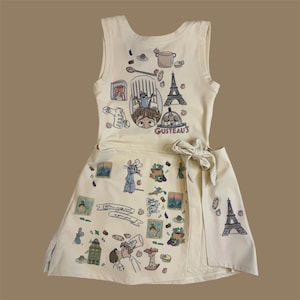 May include: Cream-colored sleeveless dress with a wrap skirt, featuring cartoon illustrations of a chef, rat, Eiffel Tower, and food items. The dress has a bow at the waist and the text "Gusteau's".
