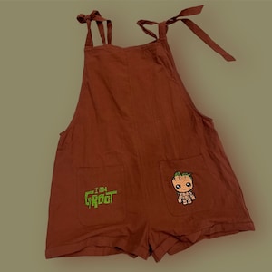 May include: Brown overall shorts with adjustable straps. The shorts have two pockets, one with an embroidered "I am Groot" design and the other with an embroidered image of Groot.
