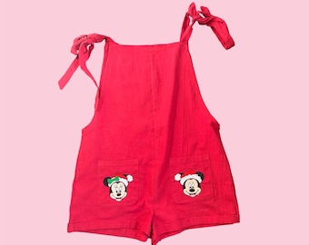 May include: Red overall shorts with adjustable straps. The shorts feature two embroidered images of Mickey Mouse wearing a Santa hat.
