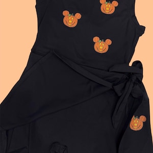 May include: Black romper with a wrap skirt and tie. The sleeveless top is adorned with three embroidered orange pumpkin Mickey Mouse designs. A fourth pumpkin design is on the wrap skirt. The romper is made of a soft, dark fabric.
