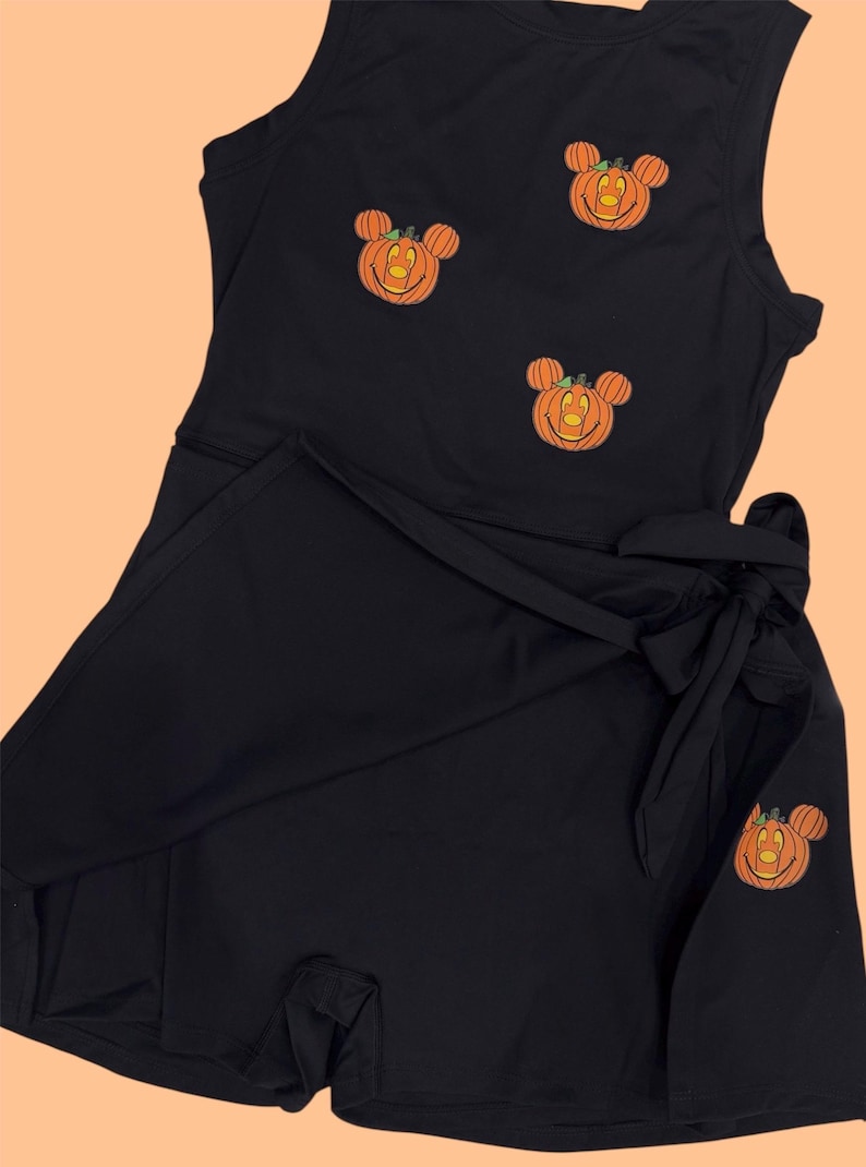 May include: Black romper with a wrap skirt and tie. The sleeveless top is adorned with three embroidered orange pumpkin Mickey Mouse designs. A fourth pumpkin design is on the wrap skirt. The romper is made of a soft, dark fabric.