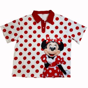 May include: White polo shirt with red polka dots and a red collar. The shirt features a graphic of Minnie Mouse in a red polka dot dress. The shirt has short sleeves and a button-up collar.