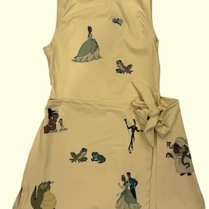 May include: A sleeveless, light yellow dress featuring cartoon illustrations of characters. The dress has a wrap-style skirt with a bow at the waist. The illustrations include a princess, frogs, and an alligator.