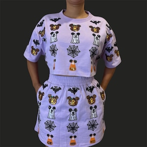 May include: A lavender two-piece outfit featuring a short-sleeved crop top and a matching skirt. The set is adorned with sequined Halloween-themed designs, including bats, jack-o'-lanterns, Mickey Mouse-like characters, ghosts, spiderwebs, and candy corn.