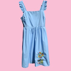 May include: A light blue dress with a house and balloons embroidered on the skirt. The dress has ruffled straps.