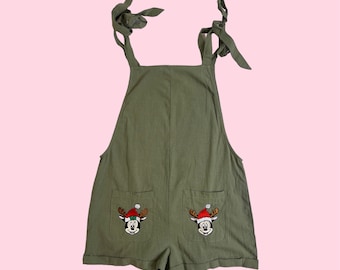 May include: Green overall shorts with adjustable straps and two pockets. Each pocket features an embroidered Mickey Mouse wearing a Santa hat and reindeer antlers.