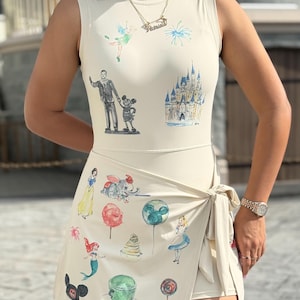 May include: Cream-colored sleeveless bodysuit and wrap skirt set featuring watercolor illustrations of Disney characters and icons. The bodysuit has images of Tinkerbell, a castle, and Mickey Mouse. The skirt has images of Dumbo, Ariel, and Alice in Wonderland.