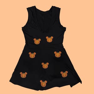 May include: Black sleeveless dress with a V-neckline. The dress features embroidered orange pumpkin designs with mouse ears. The dress has a fitted bodice and a flared skirt, perfect for Halloween or themed events.