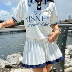 May include: A white polo shirt with a navy blue collar, short sleeves with blue trim, and the text The Disney Social Club printed in blue on the front. The shirt is paired with a matching white pleated skirt featuring a navy blue hemline.