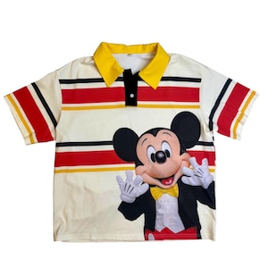 May include: Cream-colored polo shirt featuring a large graphic of Mickey Mouse. The shirt has a yellow collar, red and black stripes, and short sleeves. Mickey is wearing a tuxedo and waving.