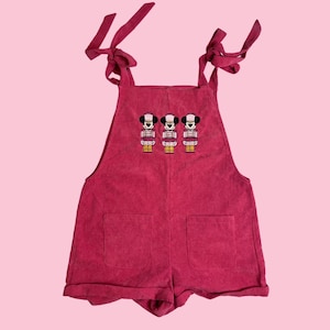 May include: A red corduroy romper with tie straps and two front pockets. The romper features an embroidered design of three Mickey Mouse figures in a row. The background is a solid pink color.