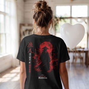 May include: A black t-shirt featuring a graphic print of a silhouetted ronin figure standing before a large red sun. The design includes vertical Japanese kanji characters, a small yin-yang symbol, and the text "The Ronin" printed in white at the bottom.