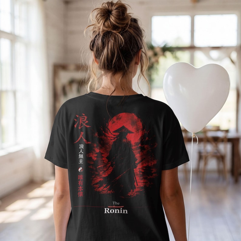 May include: A black t-shirt featuring a graphic print of a silhouetted ronin figure standing before a large red sun. The design includes vertical Japanese kanji characters, a small yin-yang symbol, and the text "The Ronin" printed in white at the bottom.